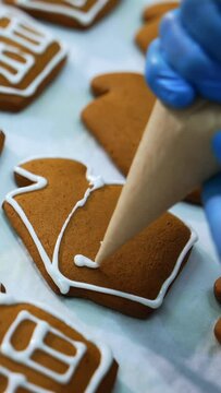 Hand in gloves decorates gingerbread houses with white icing. Close up. Pastry manufacturing at confectionery factory. Vertical video