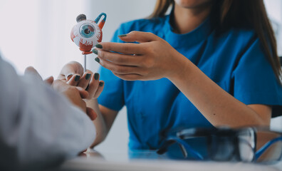 Close-up of Asian female doctor talking with elderly patient showing eyeball model and explaining...
