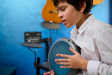 Close-up portrait teen boy with eyes closed, playing the djembe drum in a cozy home music studio,...