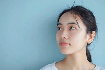 Young Asian woman with a pensive expression, gazing into the distance, blue background enhancing her serene beauty.