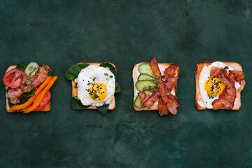  Sandwiches with fried eggs, fried ham, tomatoes, cucumbers, pepper on dark green background, top view.