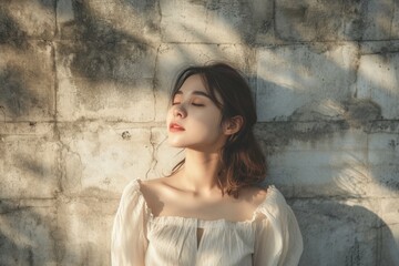 Serene young woman with eyes closed, basking in sunlight against a textured wall, conveying peace and tranquility.