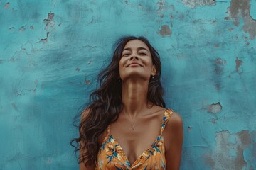 Joyful woman smiling with closed eyes against a textured blue wall, wearing a floral dress.