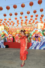 Portrait of a beautiful Asian woman in a red Chinese dress holding a fan and standing in front of a beautifully puppet lantern inside a shrine.
