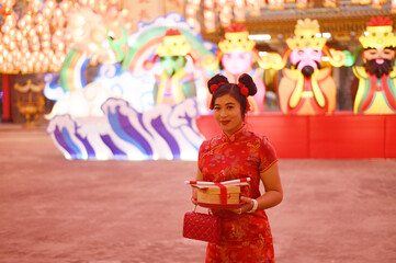 Portrait of a beautiful Asian woman in a red Chinese dress holding a gift and standing in front of a beautifully light puppet lantern inside a shrine.