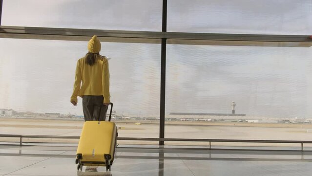 Woman with travel suitcase at airport approaches window and looks at plane taking off in anticipation of flight.
