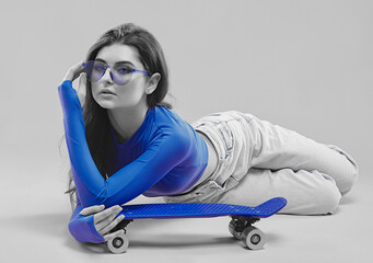A young beautiful girl in a blue jacket and denim pants with a mini skateboard on a pink background.