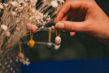 A woman hangs a tiny decorative Easter egg on a bouquet of dry white flowers in the interior....