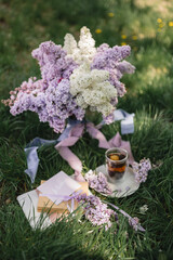 Serving on a green lawn: a lilac in a vase, a floral Chinese natural tea brewed in a glass cup, a card and purple ribbons. Tea party on a picnic. Romance