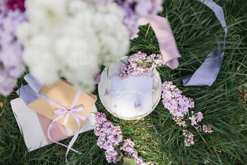 A greeting card with the inscription "Bonjour" lies on the grass, lilac flowers and a purple silk ribbon. Top view © Olga