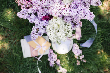 A greeting card with the inscription "Bonjour" lies on the grass, near a bouquet of purple, white and soft blue lilac and a purple silk ribbon. Decor © Olga