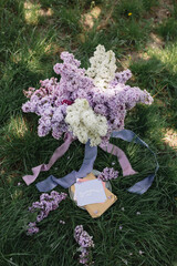 A greeting card with the inscription "Bonjour" lies on the grass, near a bouquet of purple, white and soft blue lilac and a purple silk ribbon. Decor © Olga