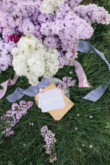 A greeting card with the inscription "Bonjour" lies on the grass, lilac flowers and a purple silk ribbon. Top view © Olga