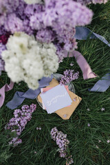 A greeting card with the inscription "Bonjour" lies on the grass, lilac flowers and a purple silk ribbon. Top view © Olga