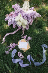 A greeting card with the inscription "Bonjour" lies on the grass, near a bouquet of purple, white and soft blue lilac and a purple silk ribbon. Decor © Olga