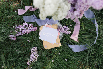 A greeting card with the inscription "Bonjour" lies on the grass, lilac flowers and a purple silk ribbon. Top view © Olga