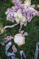 A greeting card with the inscription "Bonjour" lies on the grass, near a bouquet of purple, white and soft blue lilac and a purple silk ribbon. Decor © Olga