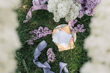 A greeting card with the inscription "Bonjour" lies on the grass, lilac flowers and a purple silk ribbon. Top view © Olga