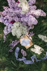 A greeting card with the inscription "Bonjour" lies on the grass, lilac flowers and a purple silk ribbon. Top view © Olga