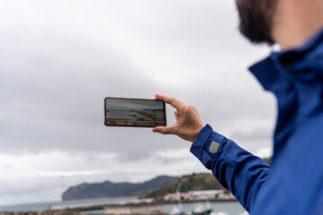 man in profile takes a picture of the coastal landscape of the Basque Country with his mobile phone on a cloudy day.
