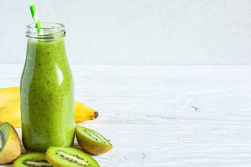 Kiwi and banana green smoothie in a glass bottle with fresh fruits on white wooden background with...