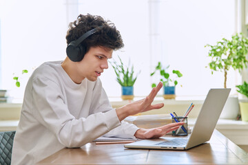 Young male college student sitting at desk, talking in web camera of laptop