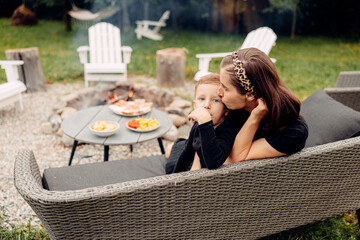 A mother kisses her son as he lays on the outdoor sofa by the barbecue., summer patio, cooking...