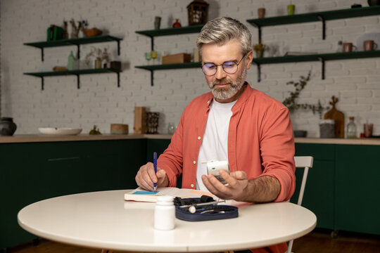 Middle aged man with glucometer checking blood sugar level at home taking notes with indicators