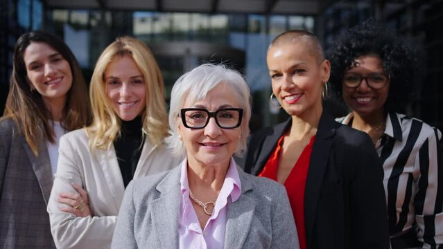 Working group formal business women of different ages looking smiling at camera together with happy expression outdoors. Mature Caucasian professional female leader posing confident in foreground 