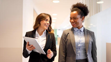 Two businesswomen going on the company meeting together, chatting and using a digital tablet on the...