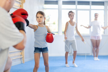 Girl and dad are boxing in gym, father helps daughter child to work out force of blow. Modern...