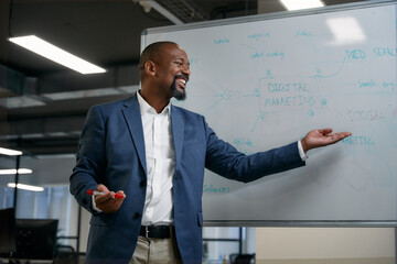 Happy mature black businessman smiling and gesturing next to whiteboard during meeting in office - 772609042