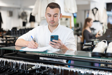 Upset man store owner makes calculations, checks documents and counts budget deficit using her phone. Male store manager is disappointed about unsatisfactory business profitability results - obrazy, fototapety, plakaty