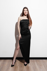A poised and elegant woman in a chic black evening dress stands confidently against a white wall,...