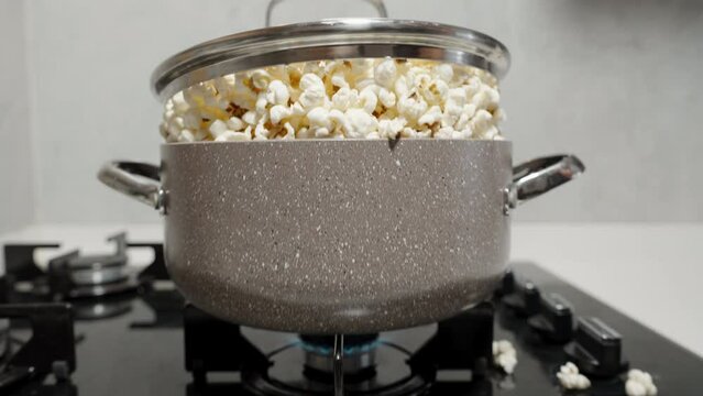 The lid of the pot rises due to the popping popcorn inside, on the gas stove, spilling over the edge.