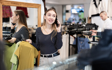 Fototapeta na wymiar Young woman in casual clothes chooses blouse and talks on phone in store