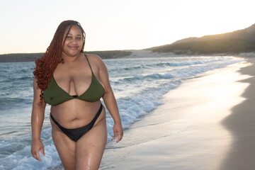 young black woman with braids walks on the beach while posing and smiling at camera. Latina plus...