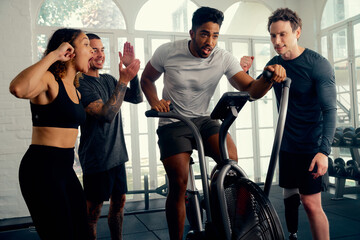 Multiracial young adults in sports clothing motivating friend cycling on exercise bike at the gym - 772634824