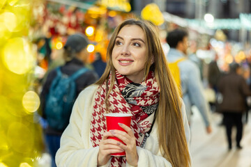 Cute happy woman with paper cup of hot drink walking at christmas fair