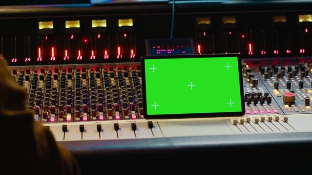 Music producer holding a tablet with greenscreen display on mixing console in control room, twisting knobs and pushing faders to edit audio recordings. Processing sounds. Camera A.