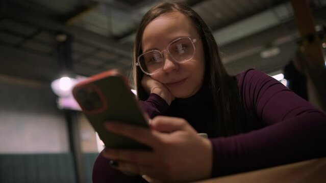 A woman comfortably sits in a warmly lit coffee shop during evening hours, sipping on fresh coffee and scroll the smartphone screen, possibly browsing the internet or checking messages.