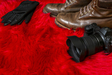 Mirrorless camera with lens hood along with brown leather boots and black gloves on a red furry surface - Powered by Adobe