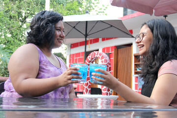Two young brunette Latin women of 20 years old drink frosty blue cocktail with chili, celebrate by drinking alcohol