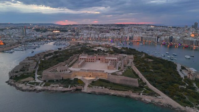 An aerial view of Maneol Island in Marsamxett Harbour, Malta. Taken at dusk with the resort town of Sliema in the background and a sunset in the distance. Flying left to right around the island