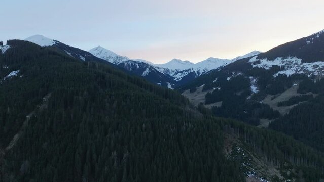 Snow-capped peaks tower over the lush pine forests of Saalbach-Hinterglemm (Austria) at dusk, aerial view