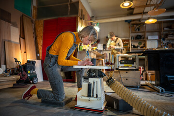 Concentrated woman carpenter works in carpentry shop, turning wooden items on lathe. Small family...