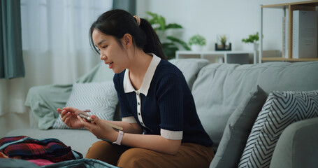 Side view of Asian teenager woman sitting on sofa making checklist of things to pack for travel, Preparation travel suitcase at home. - 772699405
