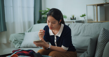 Selective focus of Asian teenager woman sitting on sofa making checklist of things to pack for travel, Preparation travel suitcase at home. - 772699418