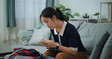 Side view of Asian teenager woman sitting on sofa making checklist of things to pack for travel, Preparation travel suitcase at home. - 772699429