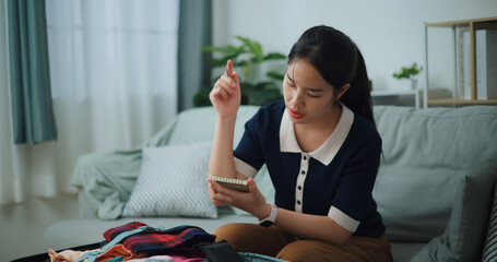Selective focus of Asian teenager woman sitting on sofa making checklist of things to pack for travel, Preparation travel suitcase at home. - 772699450